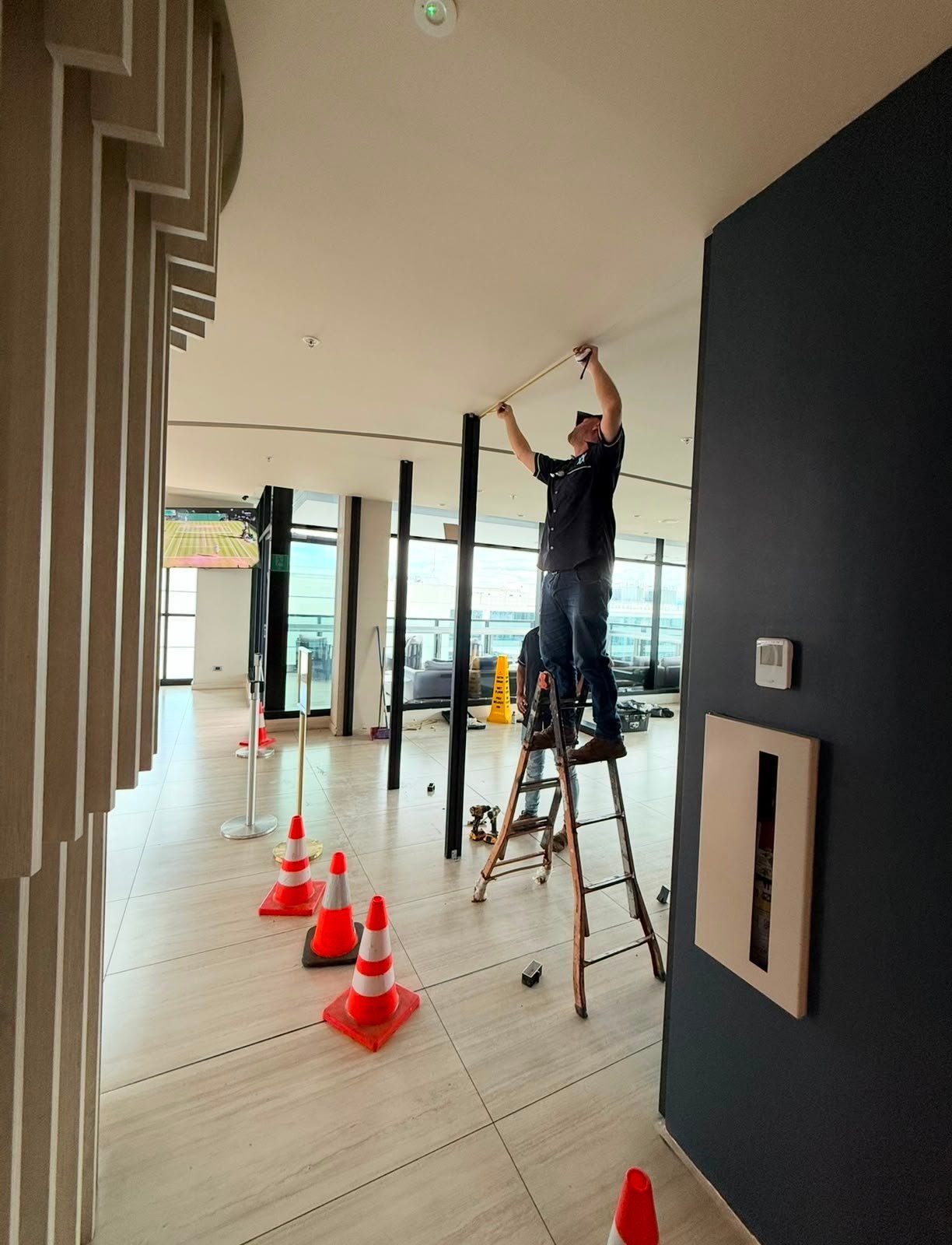 Worker on ladder installing electrical fixtures in modern commercial interior with safety cones and black accent wall