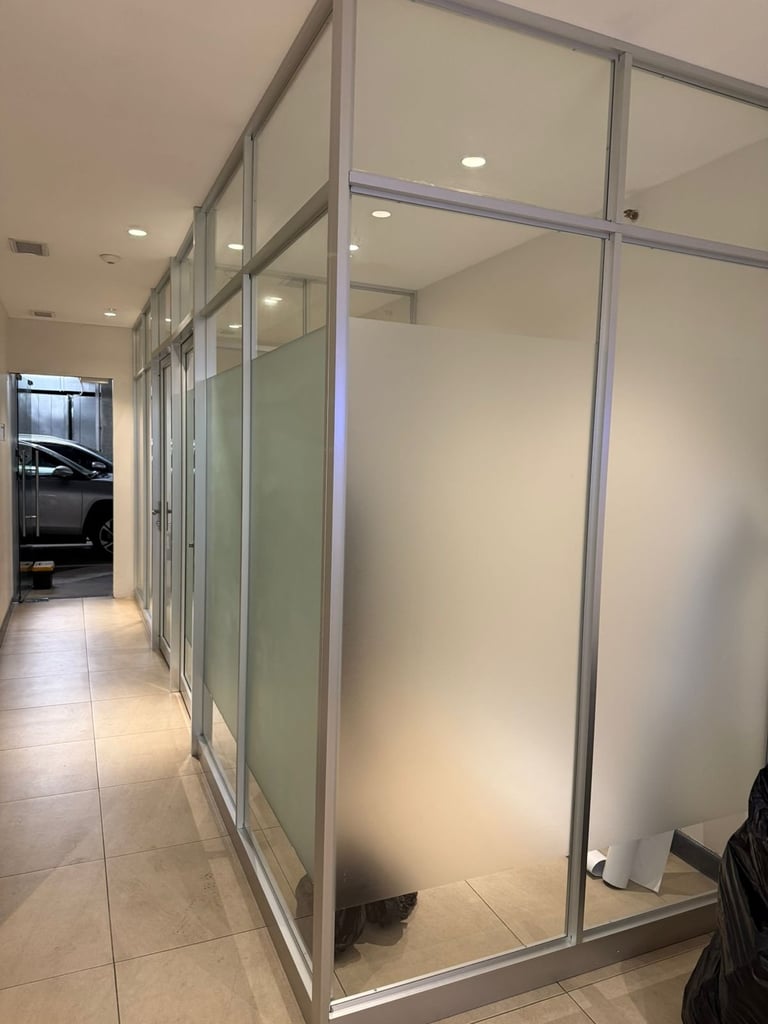 Modern office hallway with frosted glass partition walls and metal frames creating separate workspaces, with tile flooring and recessed lighting