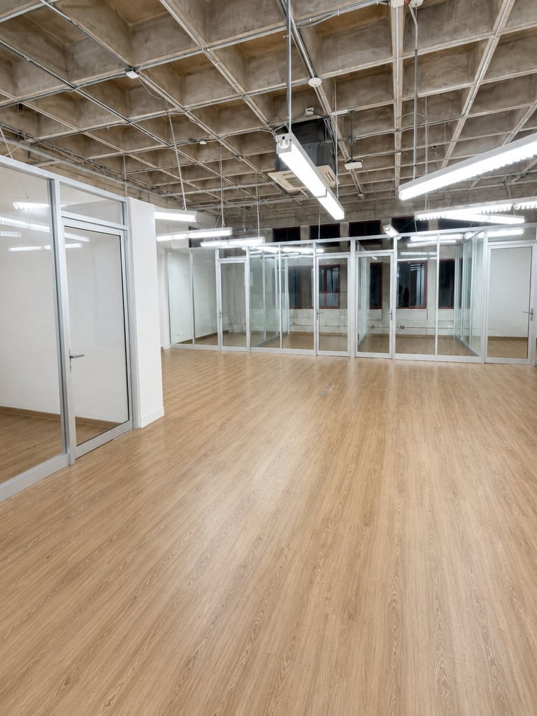 Empty modern office space with wood flooring, glass partition walls, exposed ceiling beams, and fluorescent lighting.