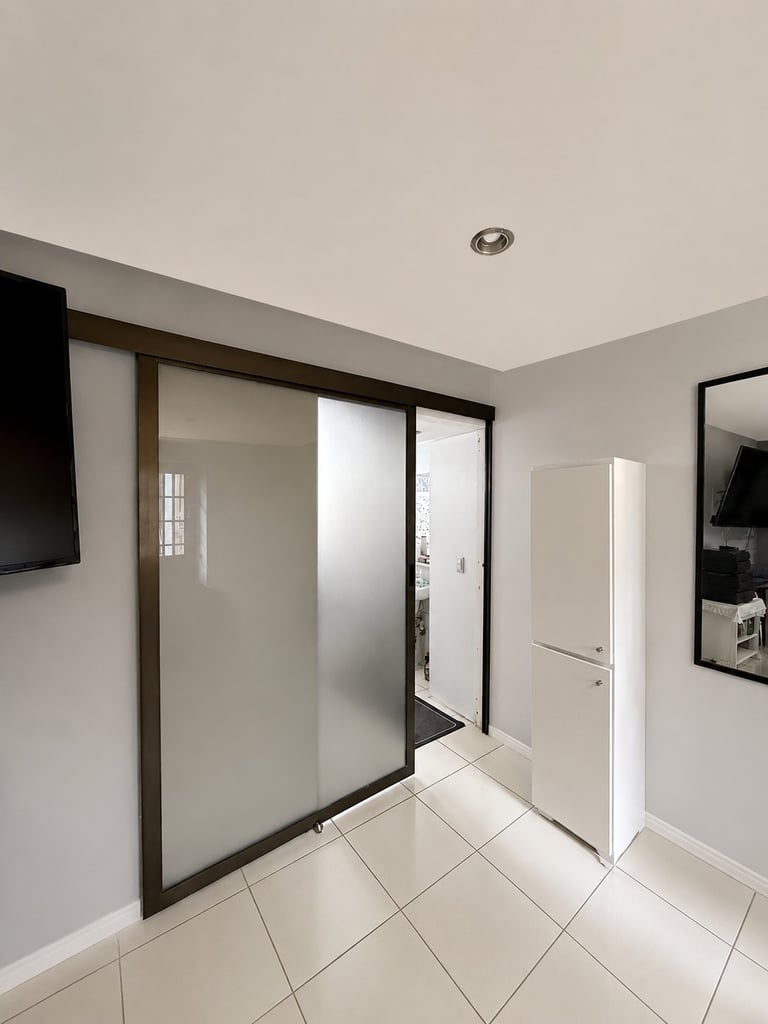 Modern bathroom entry with sliding glass doors, white tile flooring, wall-mounted TV, and recessed ceiling light