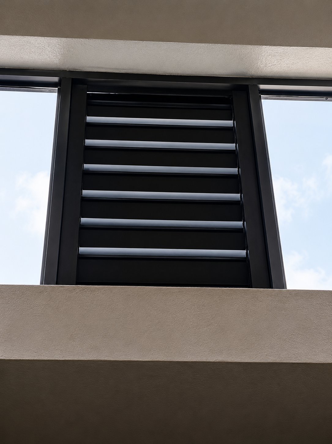 Black horizontal louvered vent window set in dark frame between light tan concrete panels on building exterior