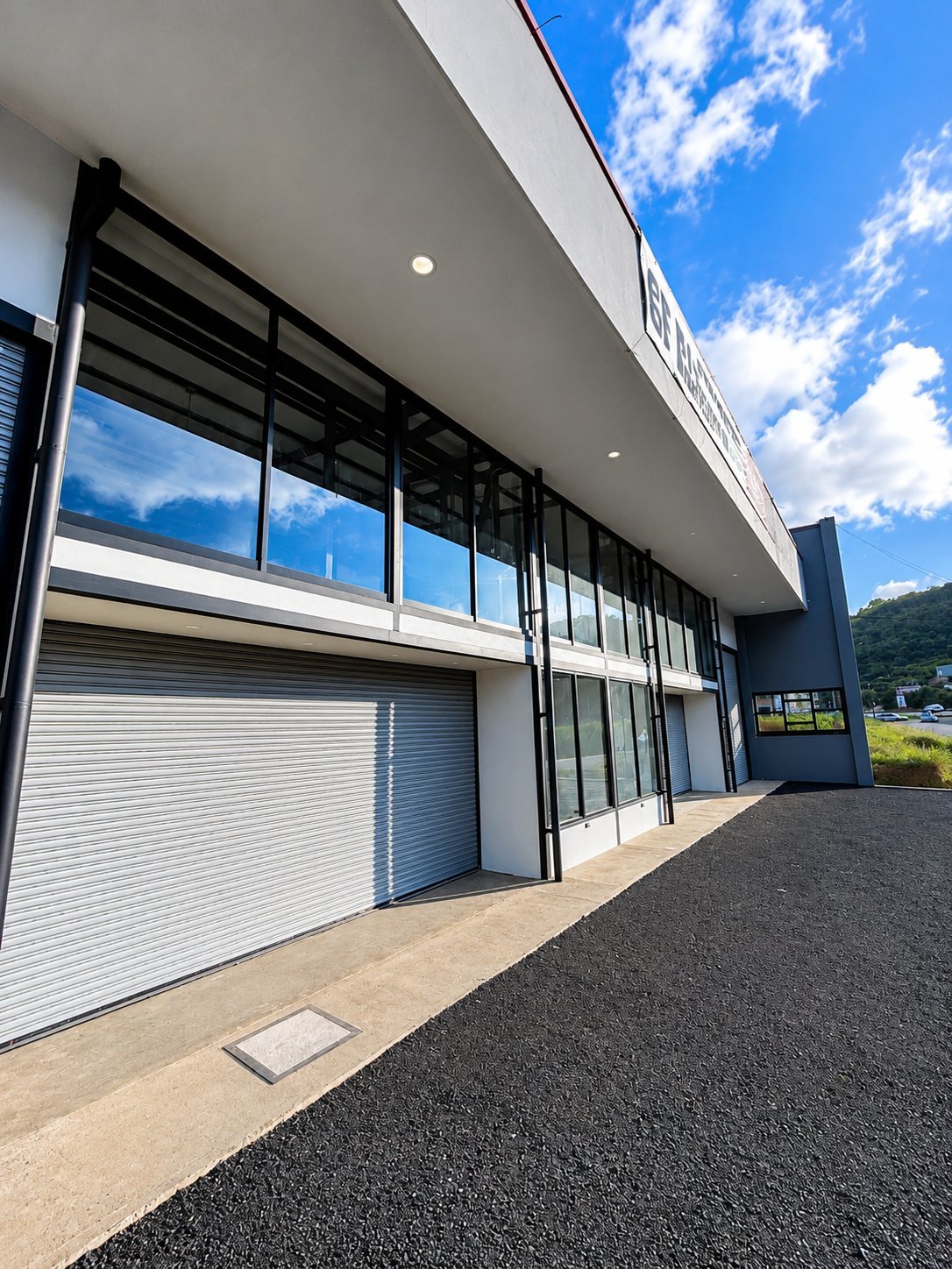 Modern commercial building exterior with white walls, black-framed glass windows, white metal garage door, and asphalt driveway under blue sky