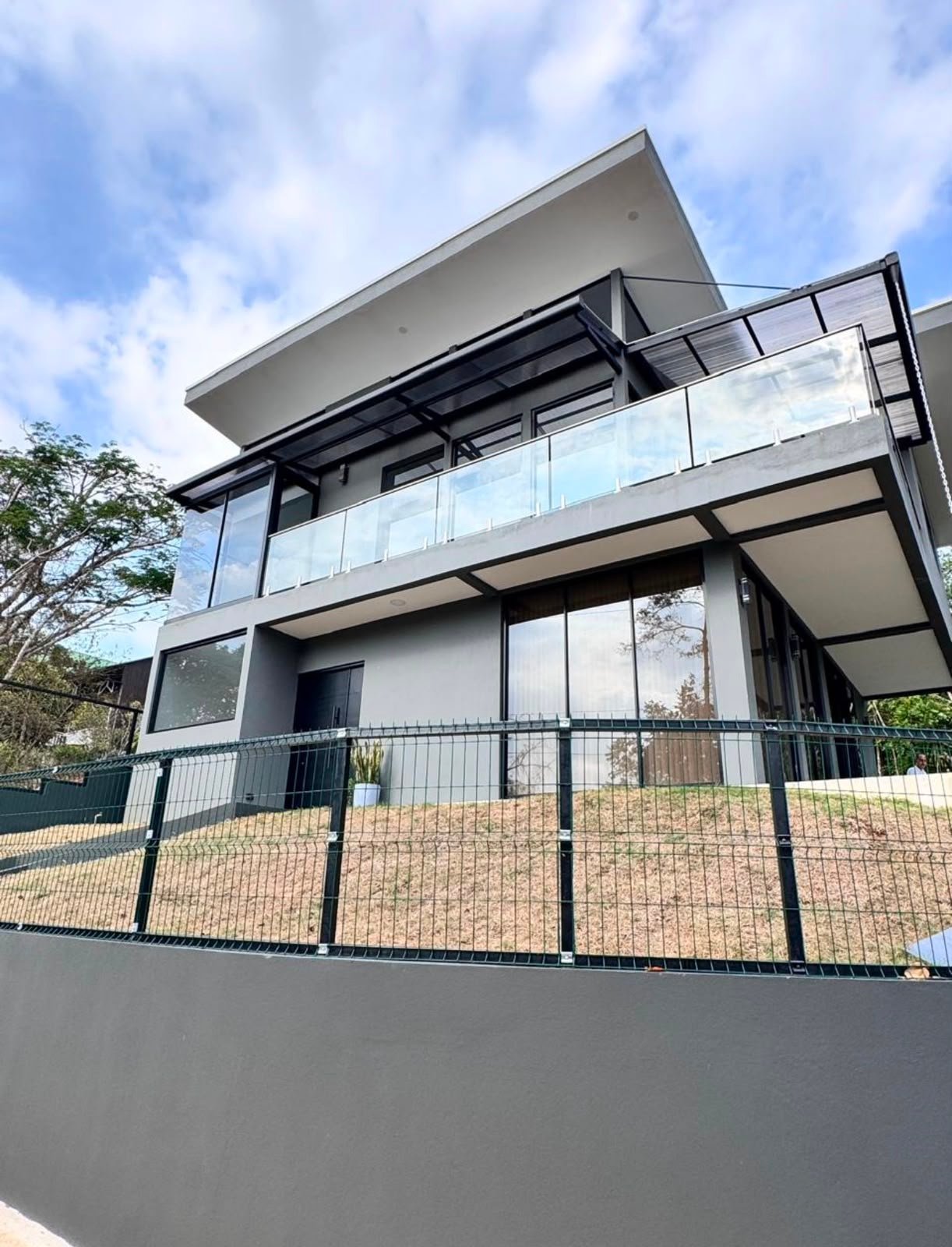Modern two-story house with white exterior, black metal railings, large glass windows, and a metal gate fence in front under blue sky
