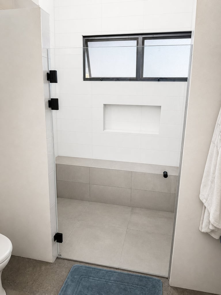 Modern frameless glass shower enclosure with black hardware and two awning windows, attached to white tiled bathroom wall