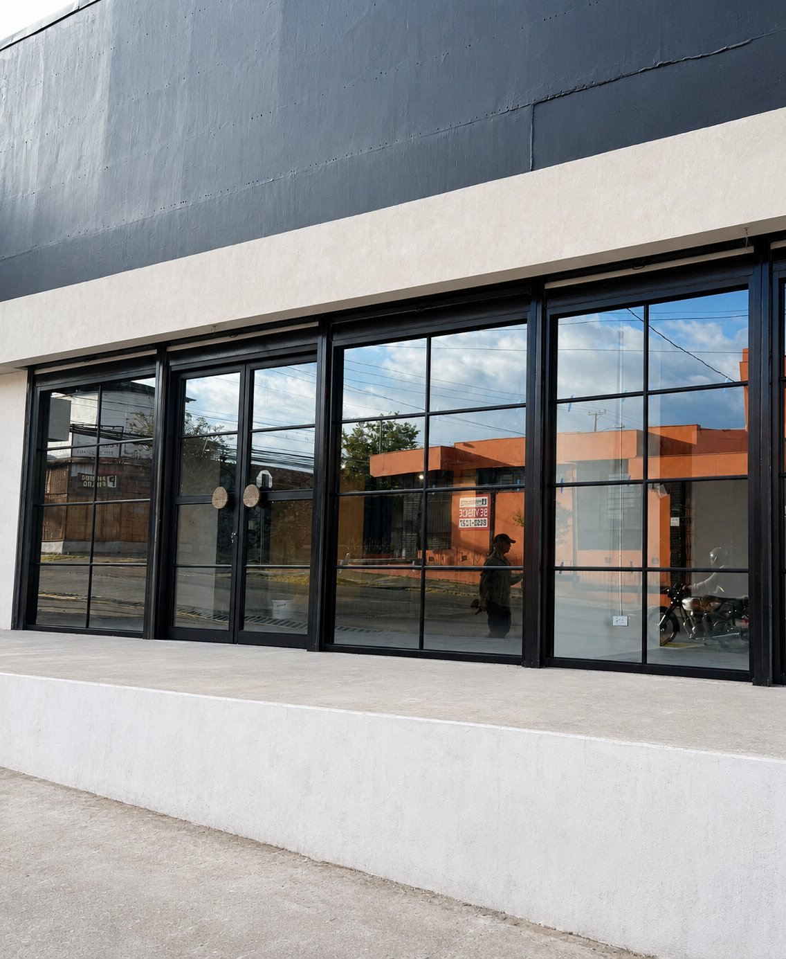 Modern storefront with black-framed glass doors and windows on white concrete facade, reflecting urban surroundings