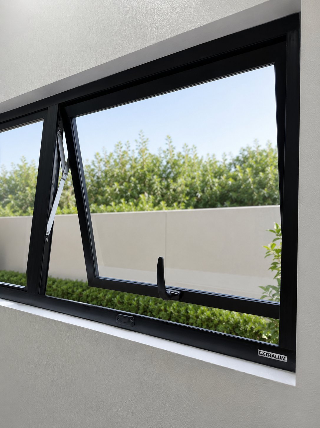 Black awning window mounted on white wall with clear view of outdoor terrace and green hedges beyond