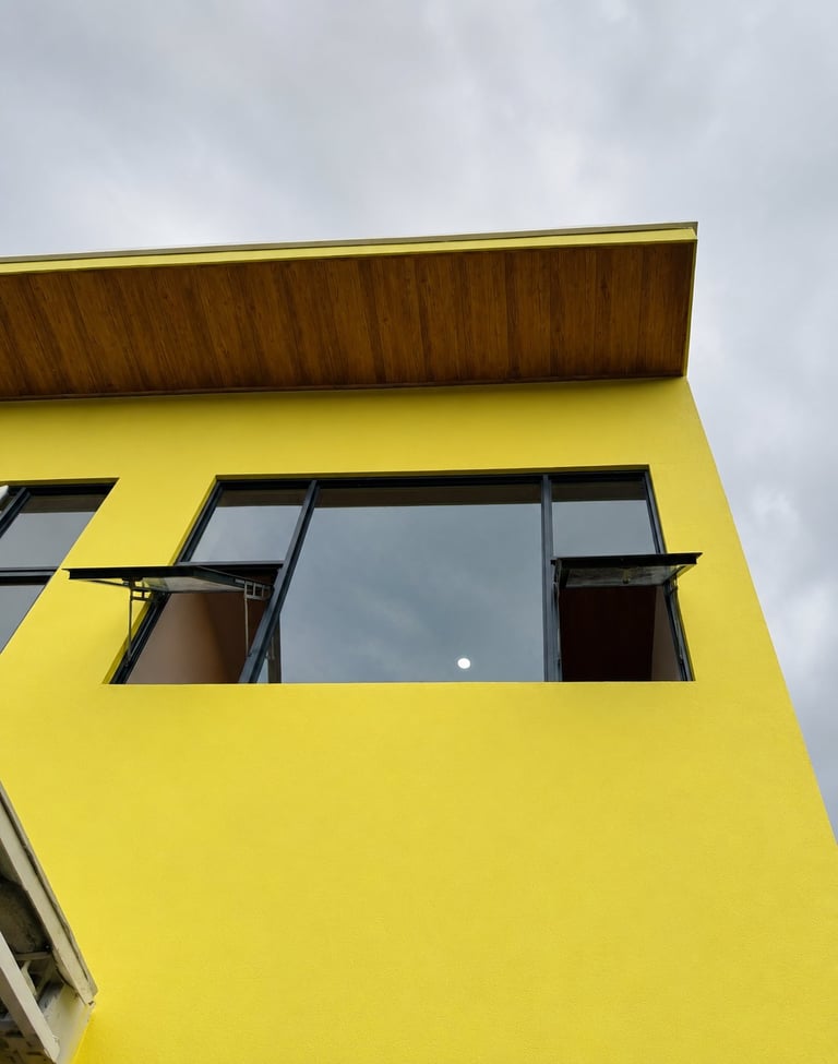 Bright yellow modern building facade with large horizontal window and wooden top trim against cloudy sky